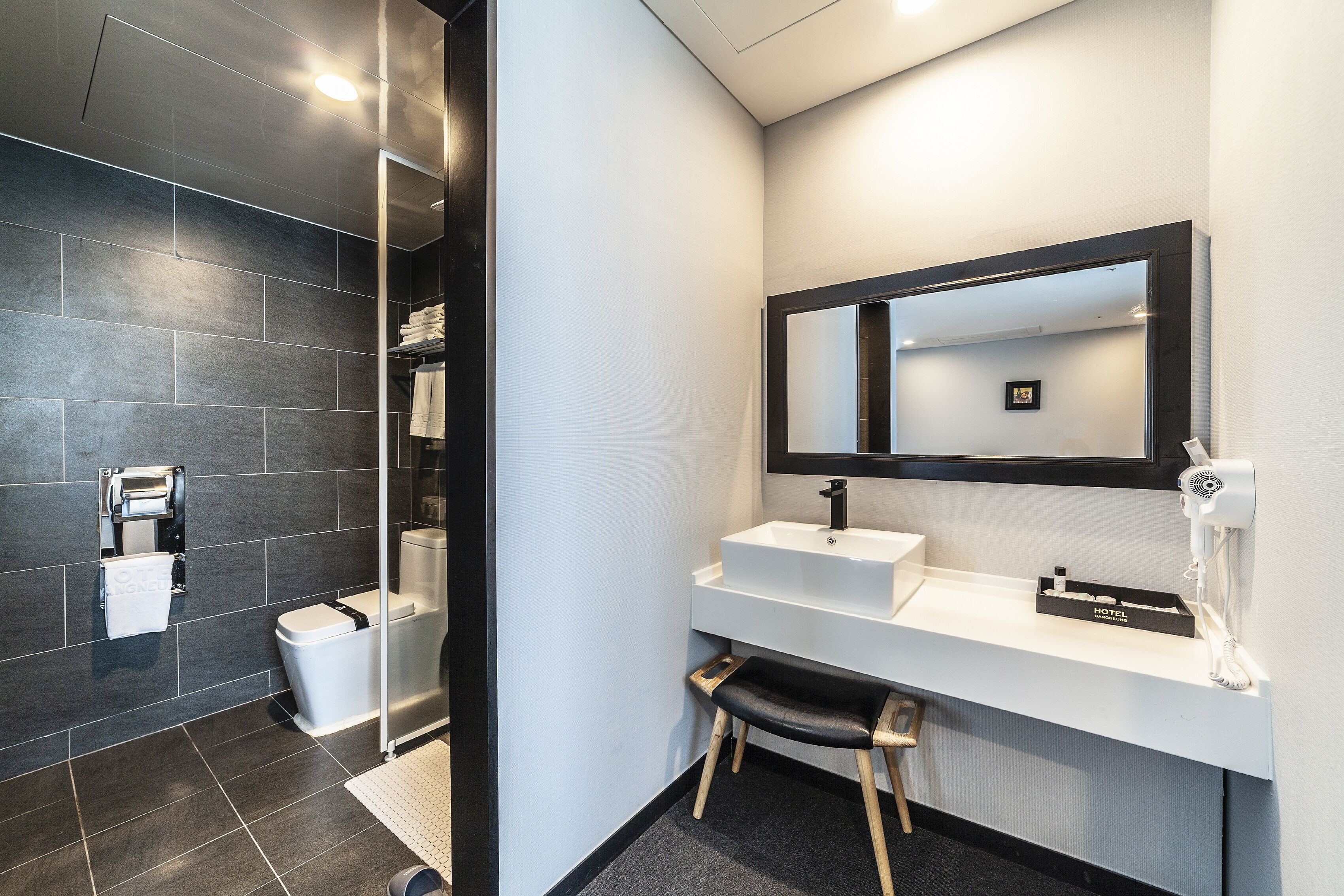 Un baño moderno con una pared de azulejos oscuros, un lavamanos blanco, un espejo y un dispensador de toallas.