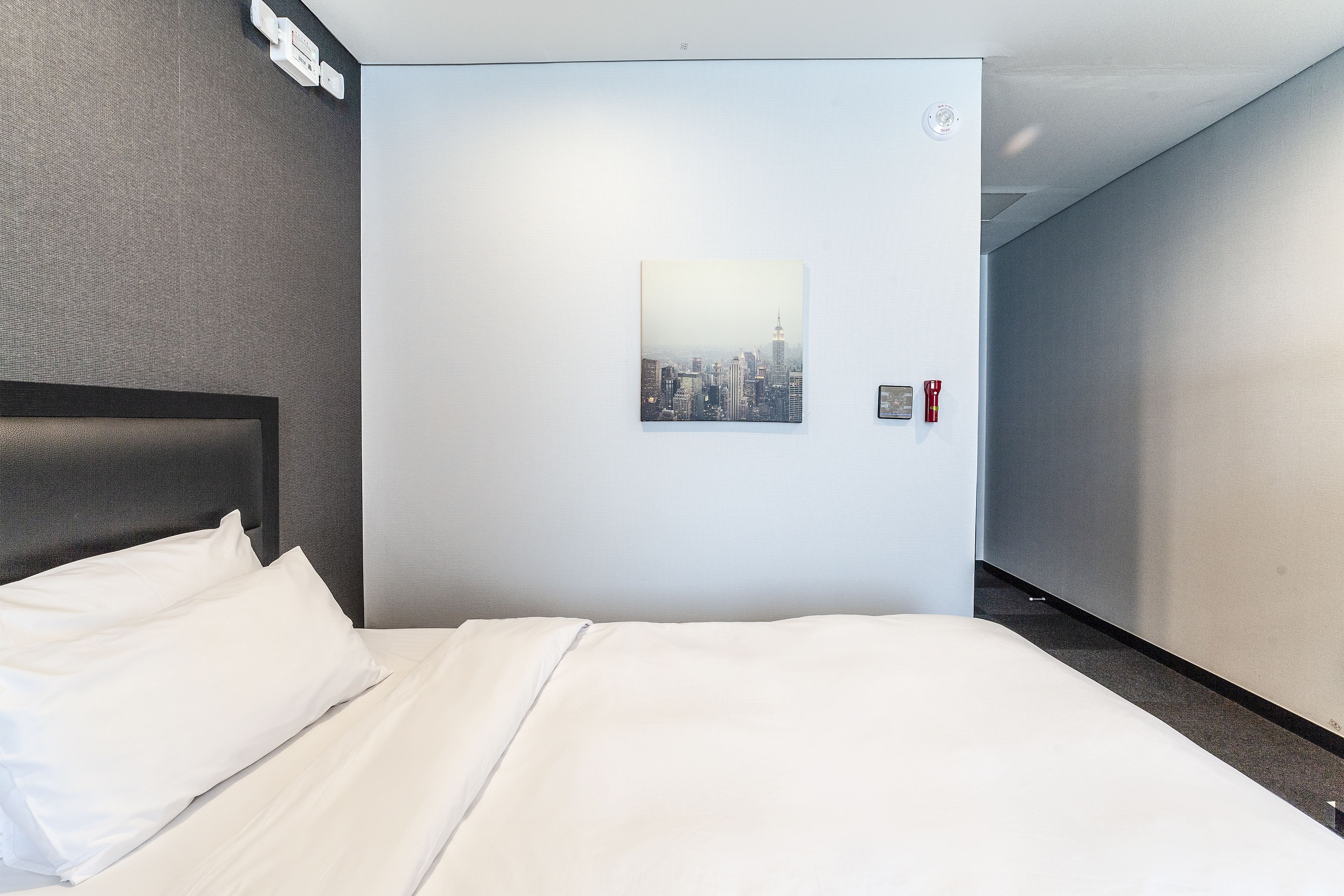 A hotel room with a bed, a cityscape picture on the wall, and a fire alarm.