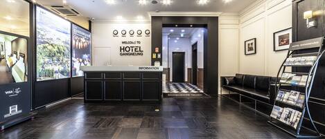 Zona de recepción de un hotel con piso de madera oscura, un mostrador de recepción, un área de espera con un sofá de cuero negro, y una pared con un cartel de bienvenida y un directorio.