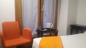 A hotel room with a bed, an orange chair, a small table with a coffee maker, and a window with curtains. - Pensión El Paseo (Pamplona)
