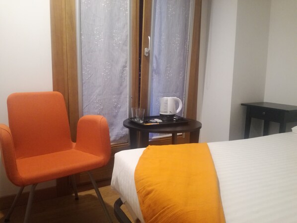 A hotel room with a bed, an orange chair, a small table with a coffee maker, and a window with curtains.