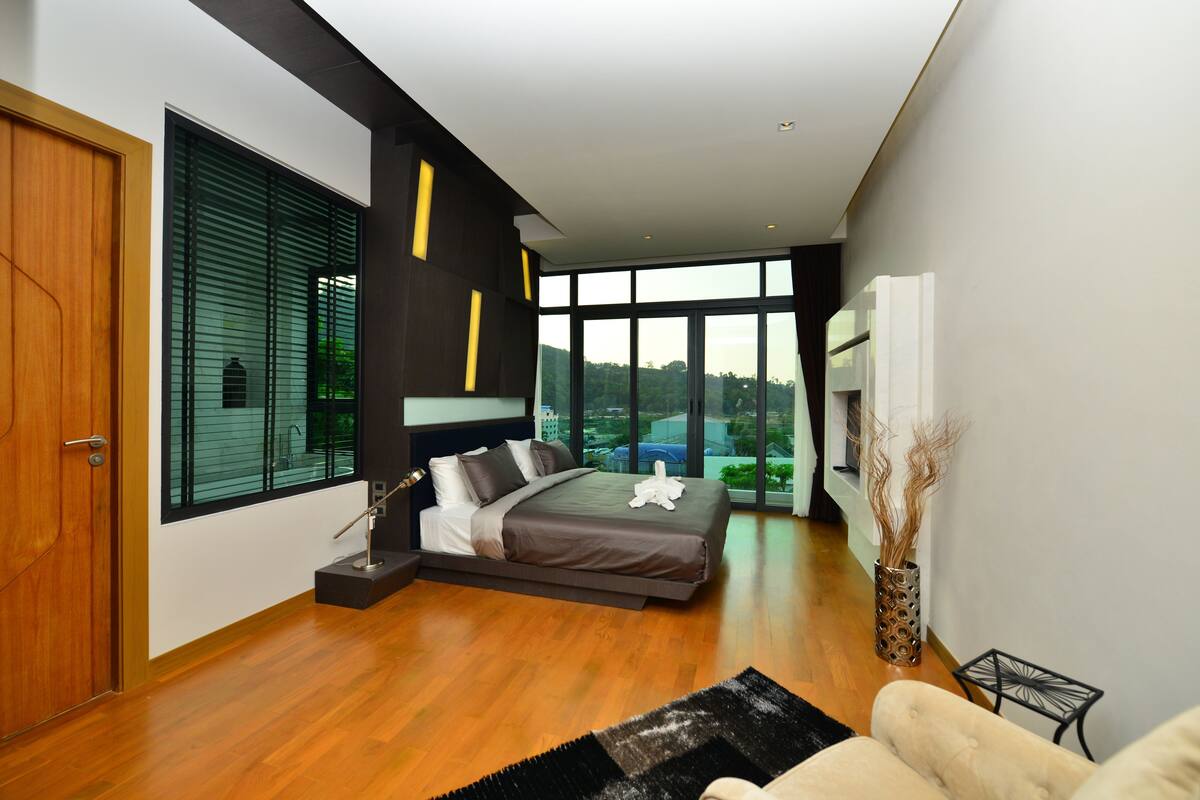 A modern bedroom with a large bed, a wooden door, and a view of greenery through large windows.