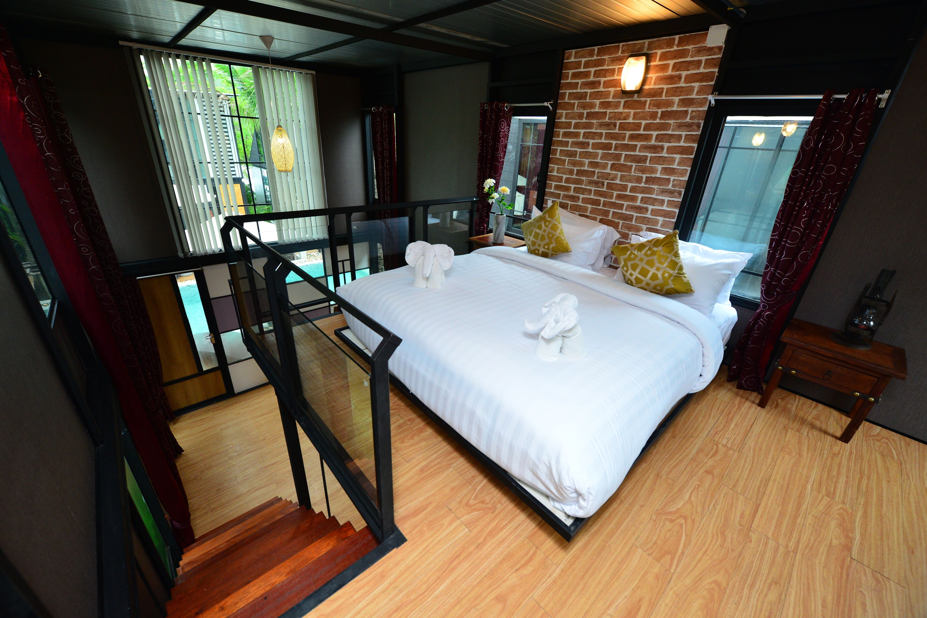 A hotel room with a large bed, a wooden floor, a brick accent wall, and a balcony with a glass railing.
