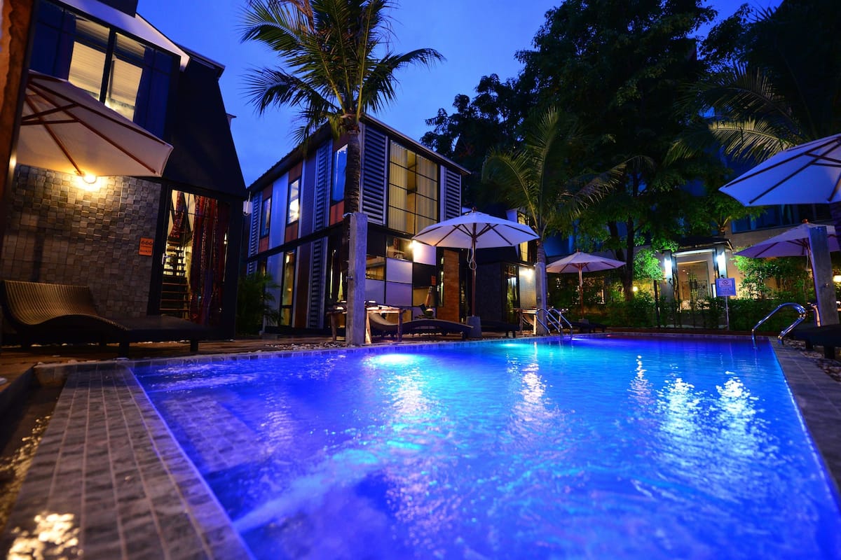 A swimming pool with a tiled edge, surrounded by lounge chairs and umbrellas, and a building with large windows and a balcony.