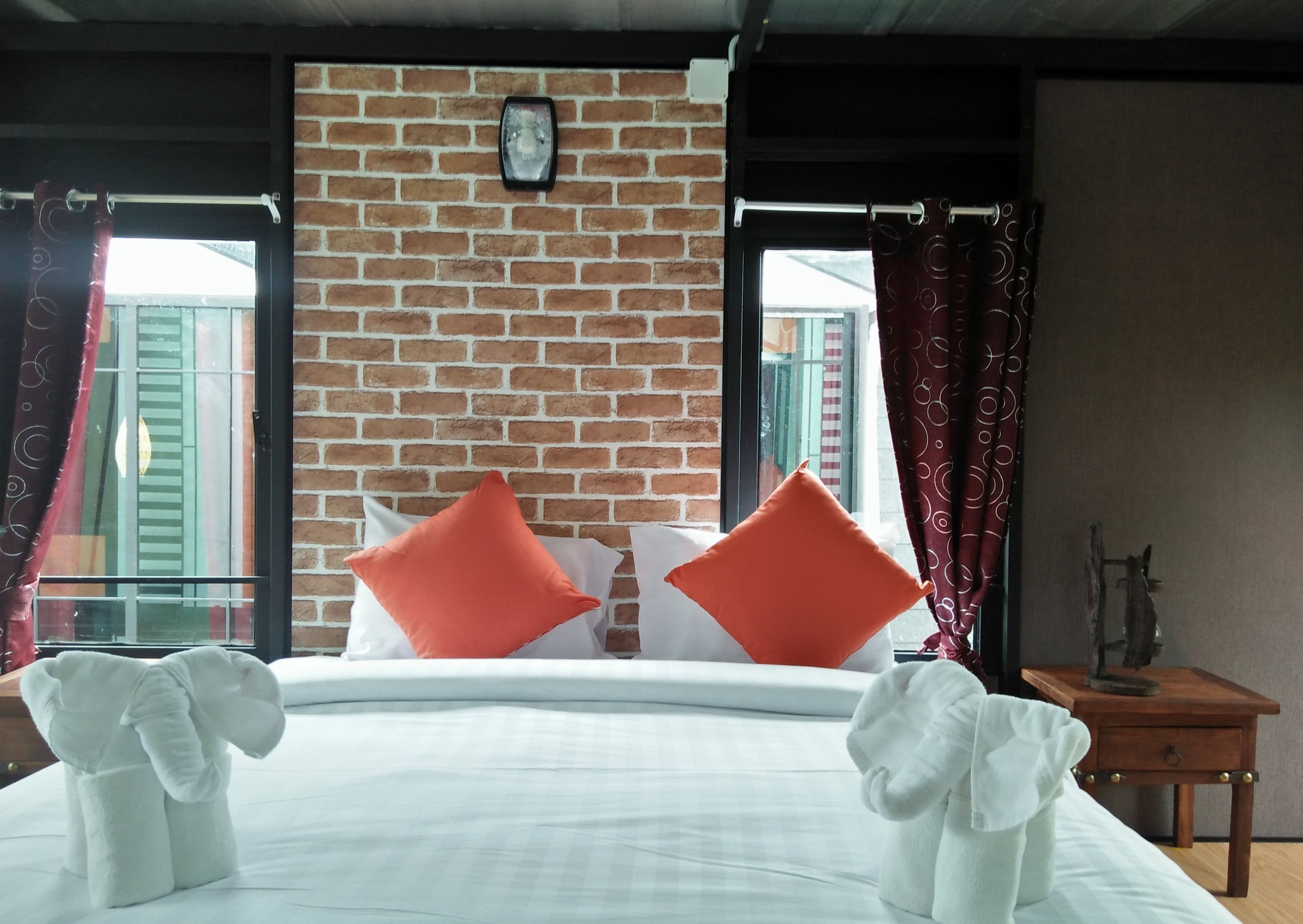 Habitación de hotel con pared de ladrillo, cama con almohadas blancas y anaranjadas, mesita de noche de madera y ventana con cortinas.