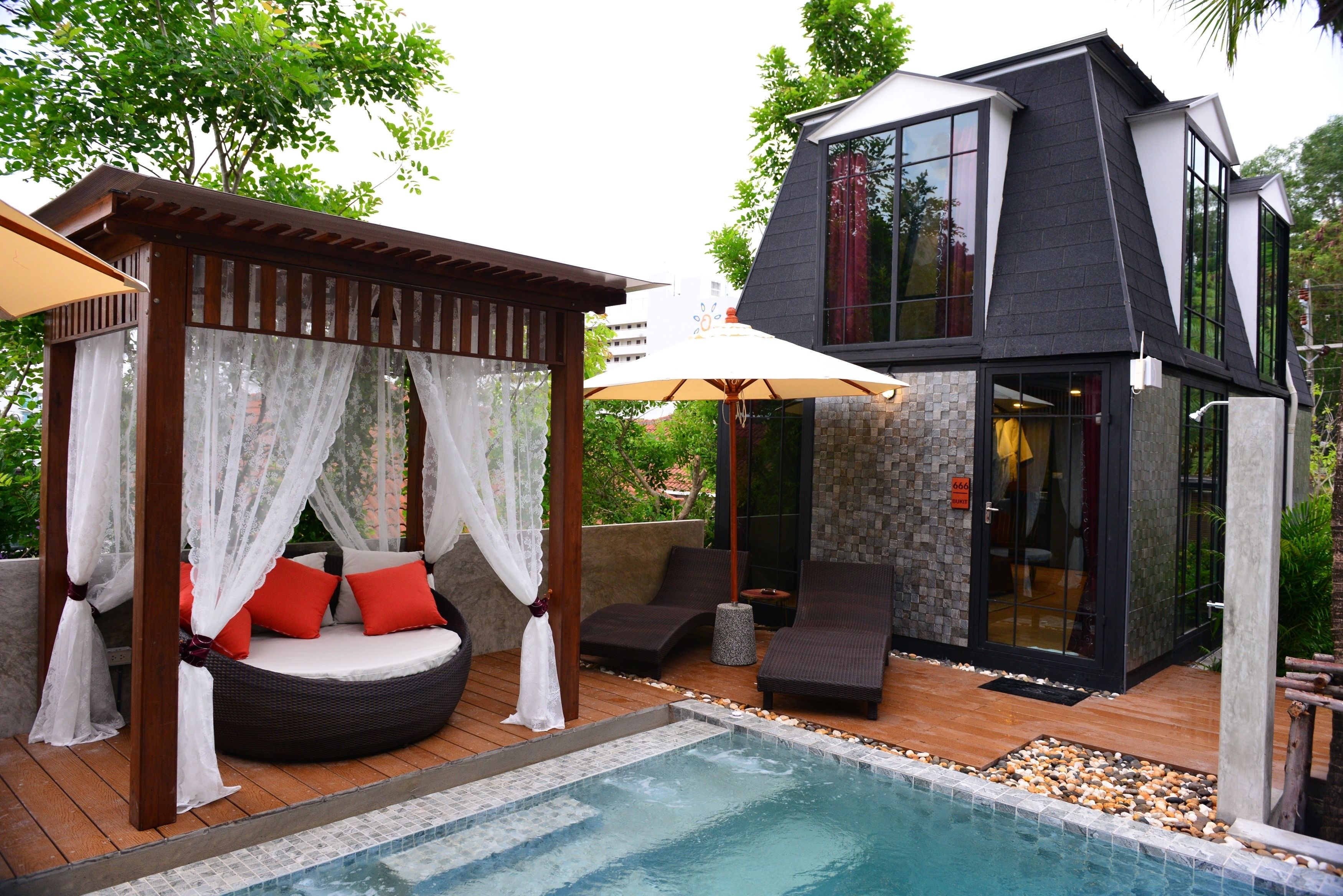 A modern outdoor pool area with a wooden deck, a hot tub, and a glass-enclosed room with a stone facade.