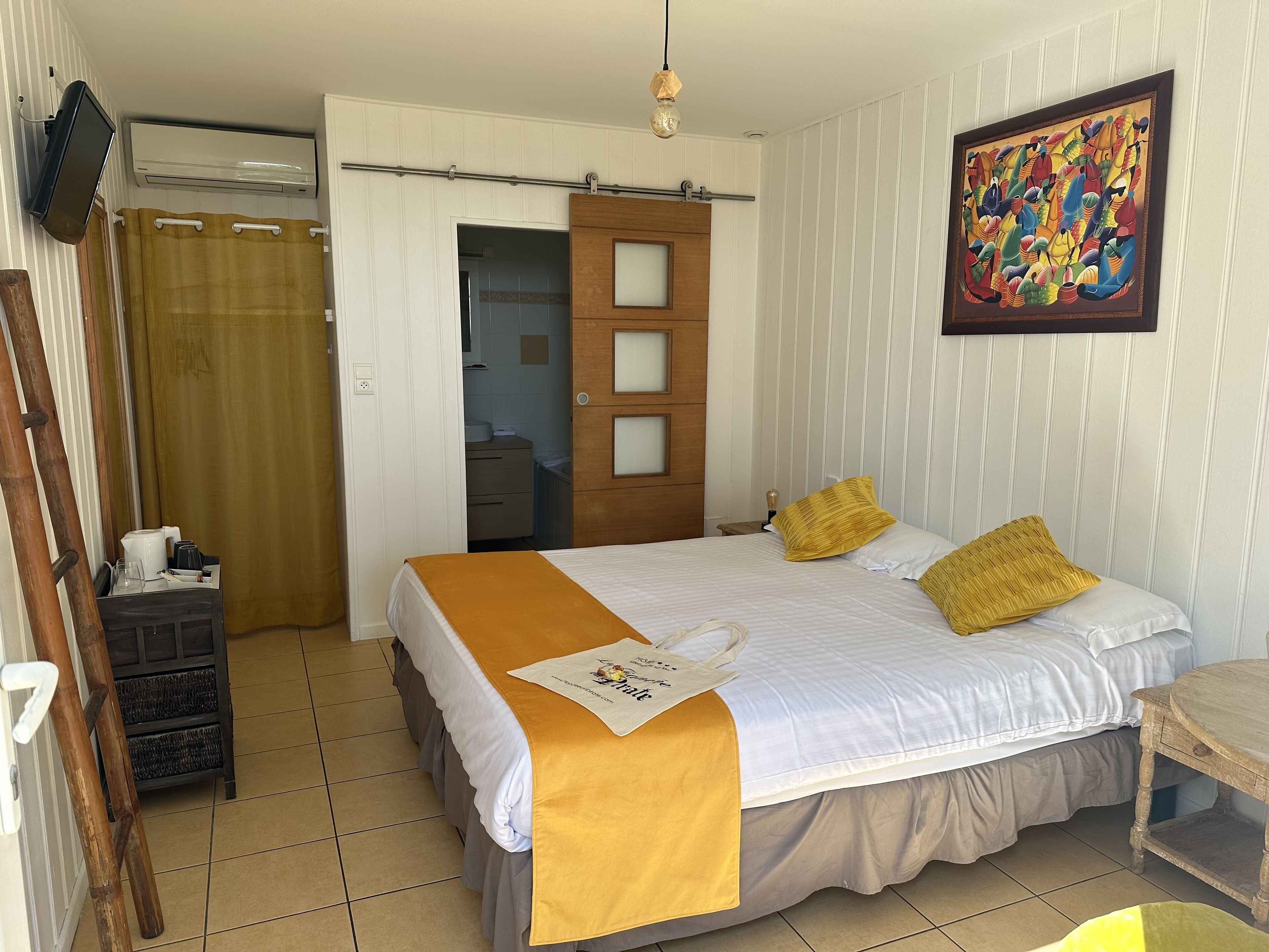 A hotel room with a bed, a wooden nightstand, a desk, a chair, a painting, and a door leading to a bathroom.