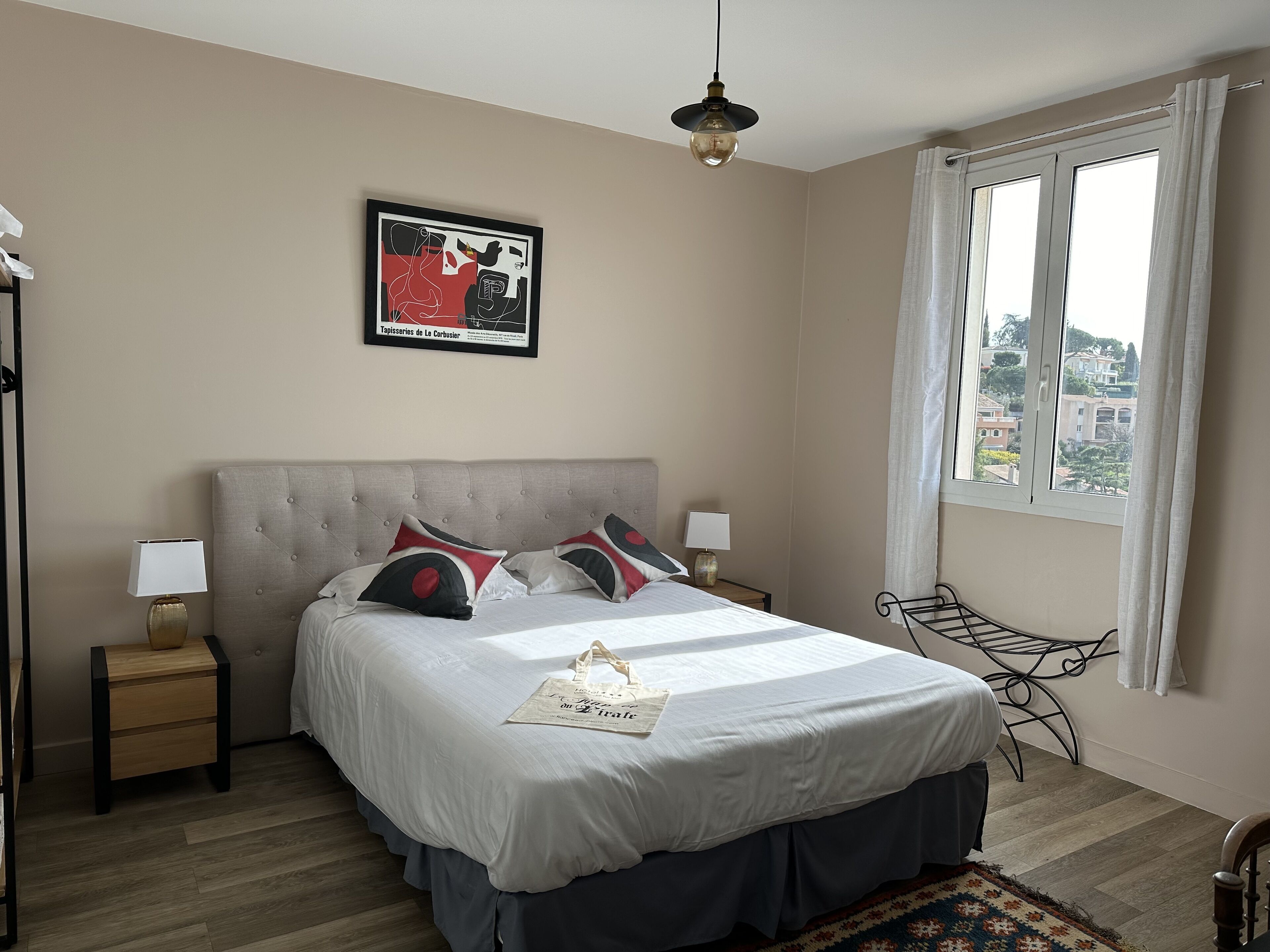 A neatly arranged bedroom with a bed, bedside tables, a hanging light fixture, a window with curtains, and a view of buildings outside.
