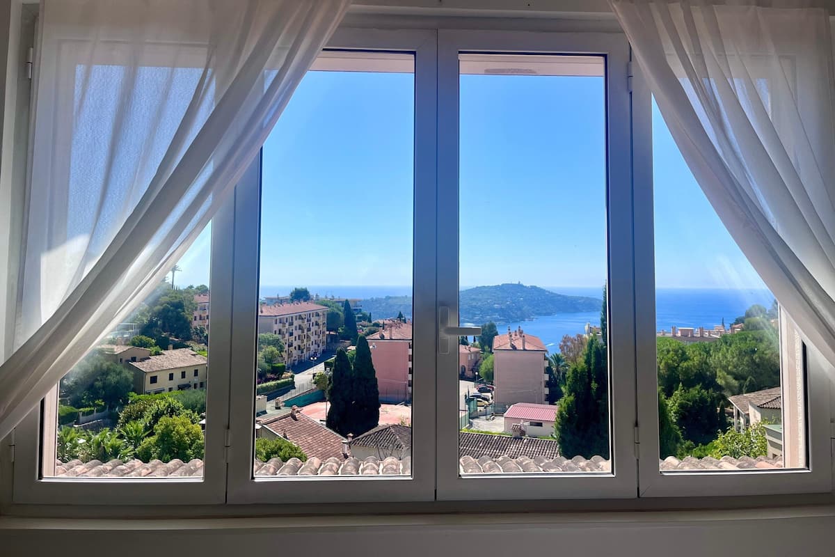A room with a large window offering a view of a coastal town and the sea.