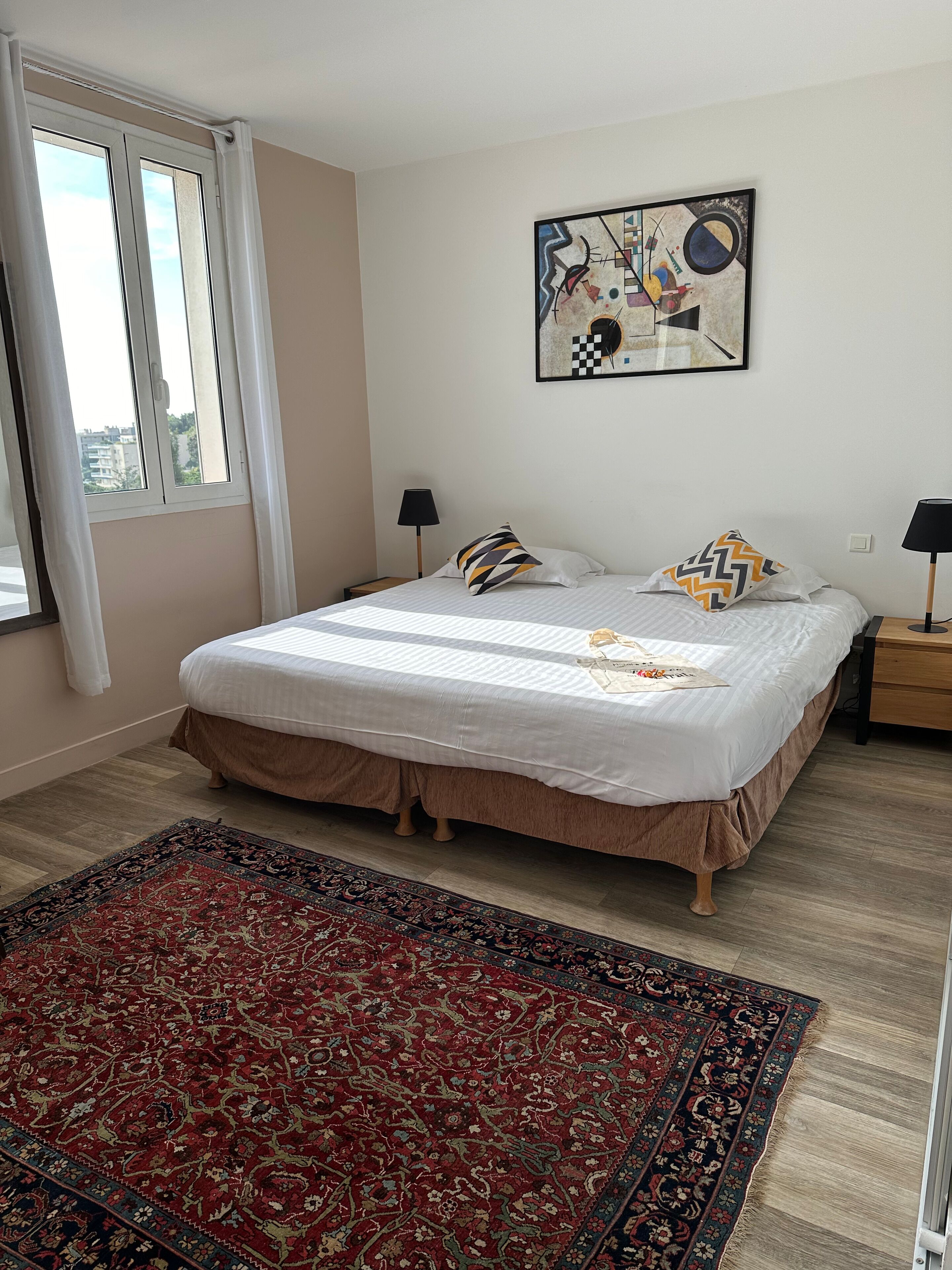 A bedroom with a large bed, a red patterned rug, a painting on the wall, and a window with curtains.