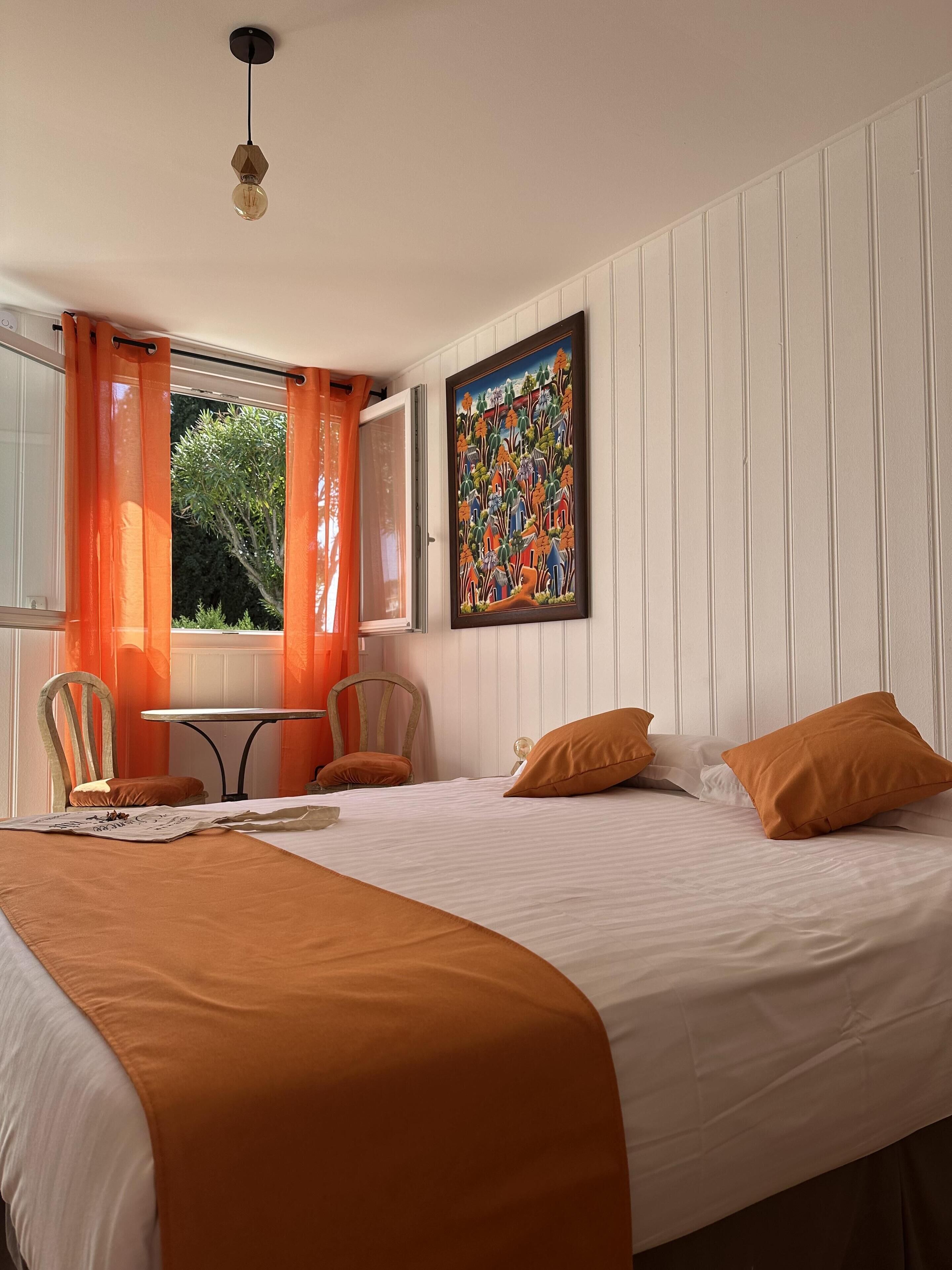 A bedroom with a bed, orange pillows, a table, chairs, and a painting on the wall.