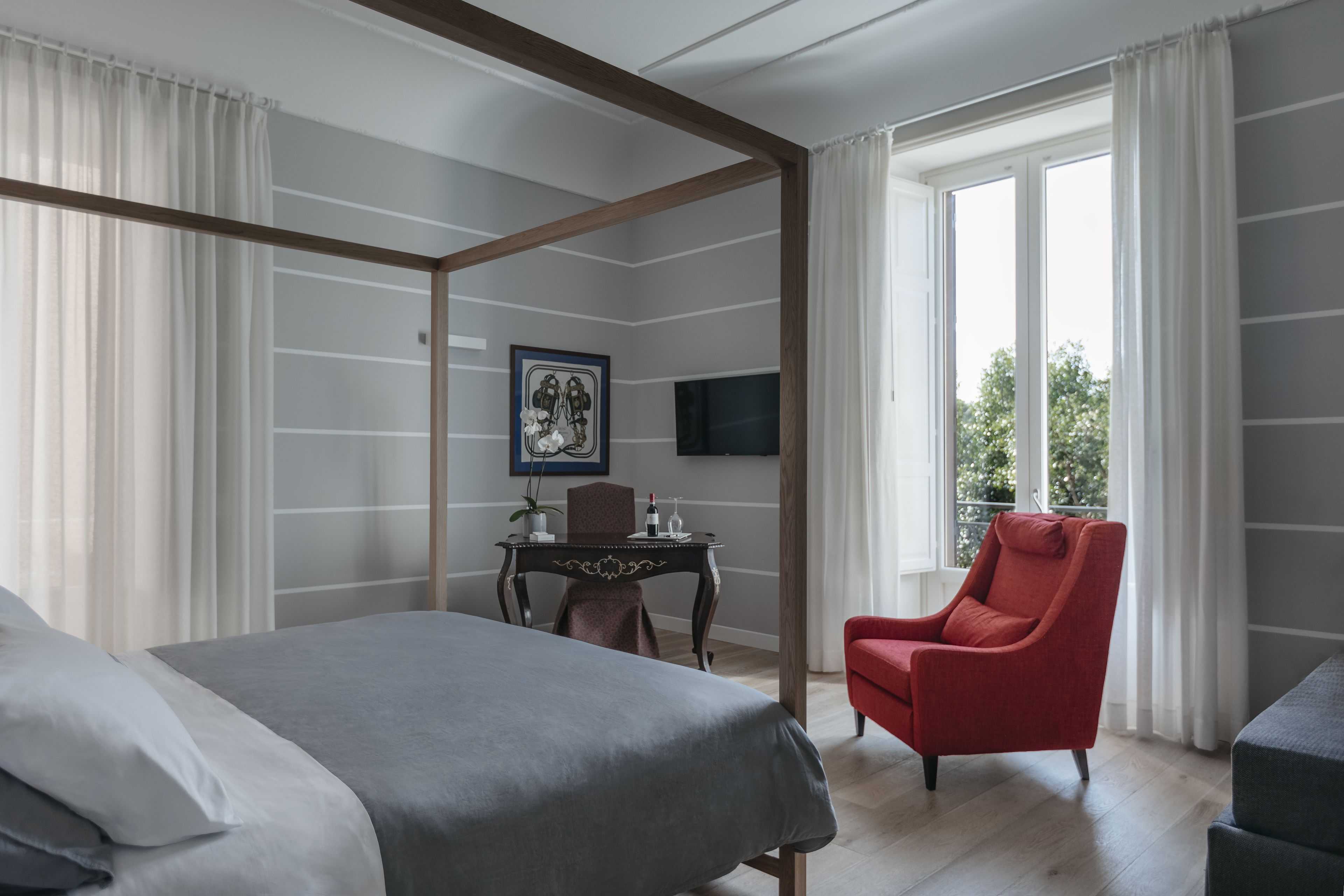 Una habitación de hotel moderna con cama con dosel, un sillón rojo, una mesita con una planta y un televisor montado en la pared.