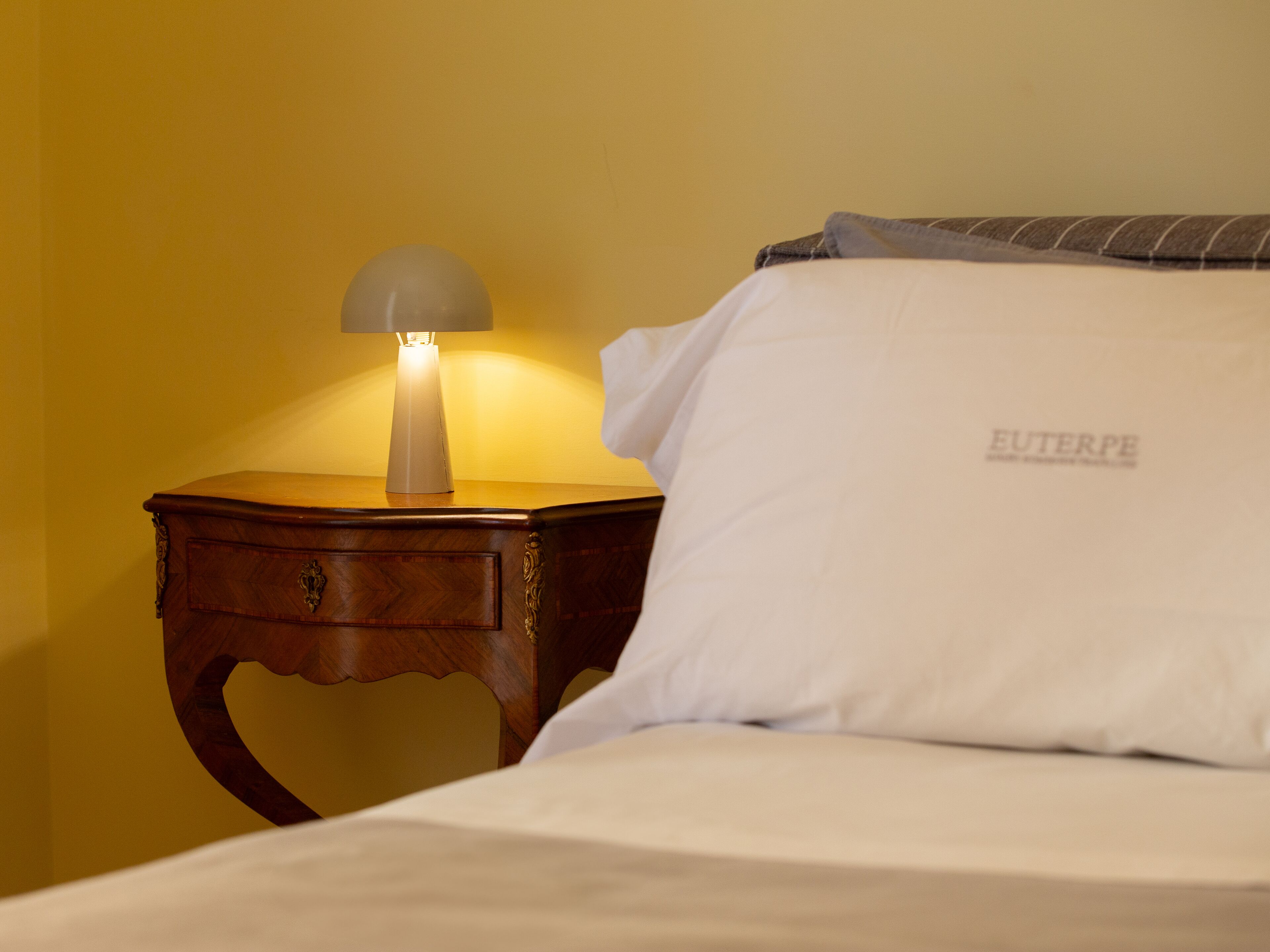 A bed with a white pillow and a wooden nightstand with a lamp.