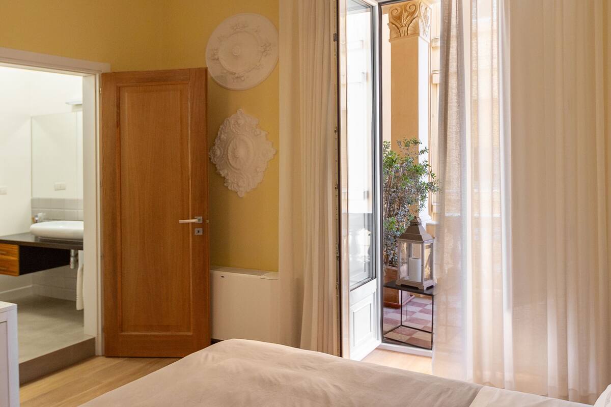 A bedroom with a large bed, a chandelier, a wooden door, and a view of the outside through a glass door.