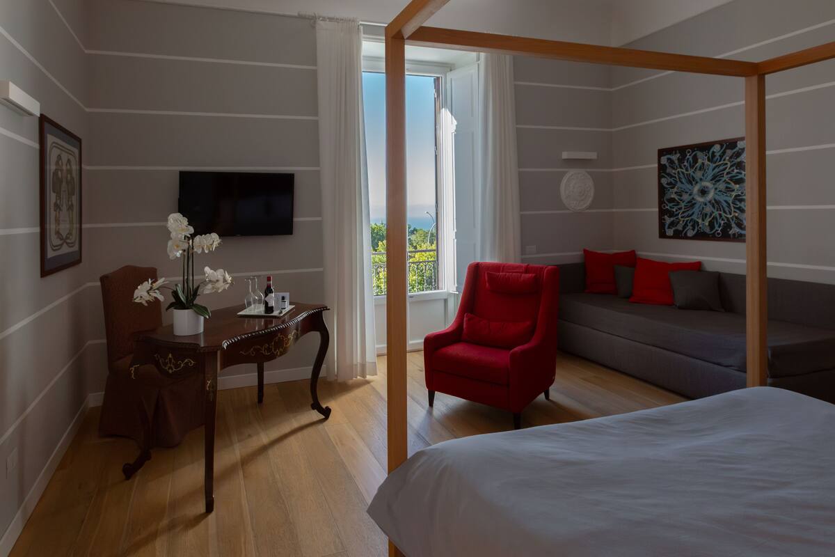 A bedroom with a four-poster bed, a red armchair, a grey sofa, a wooden desk with a vase of flowers, and a television on the wall.