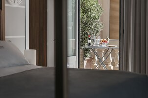 Habitación de hotel con cama, ventana que ofrece vistas a un balcón y una mesa con una botella, vasos y fruta.