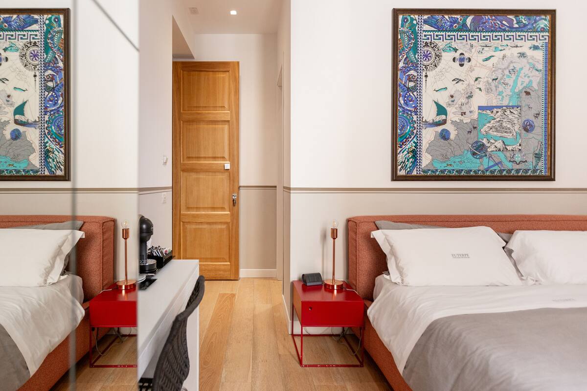 A hotel room with two beds, a wooden door, a red nightstand, a desk with a lamp, and a large map on the wall.