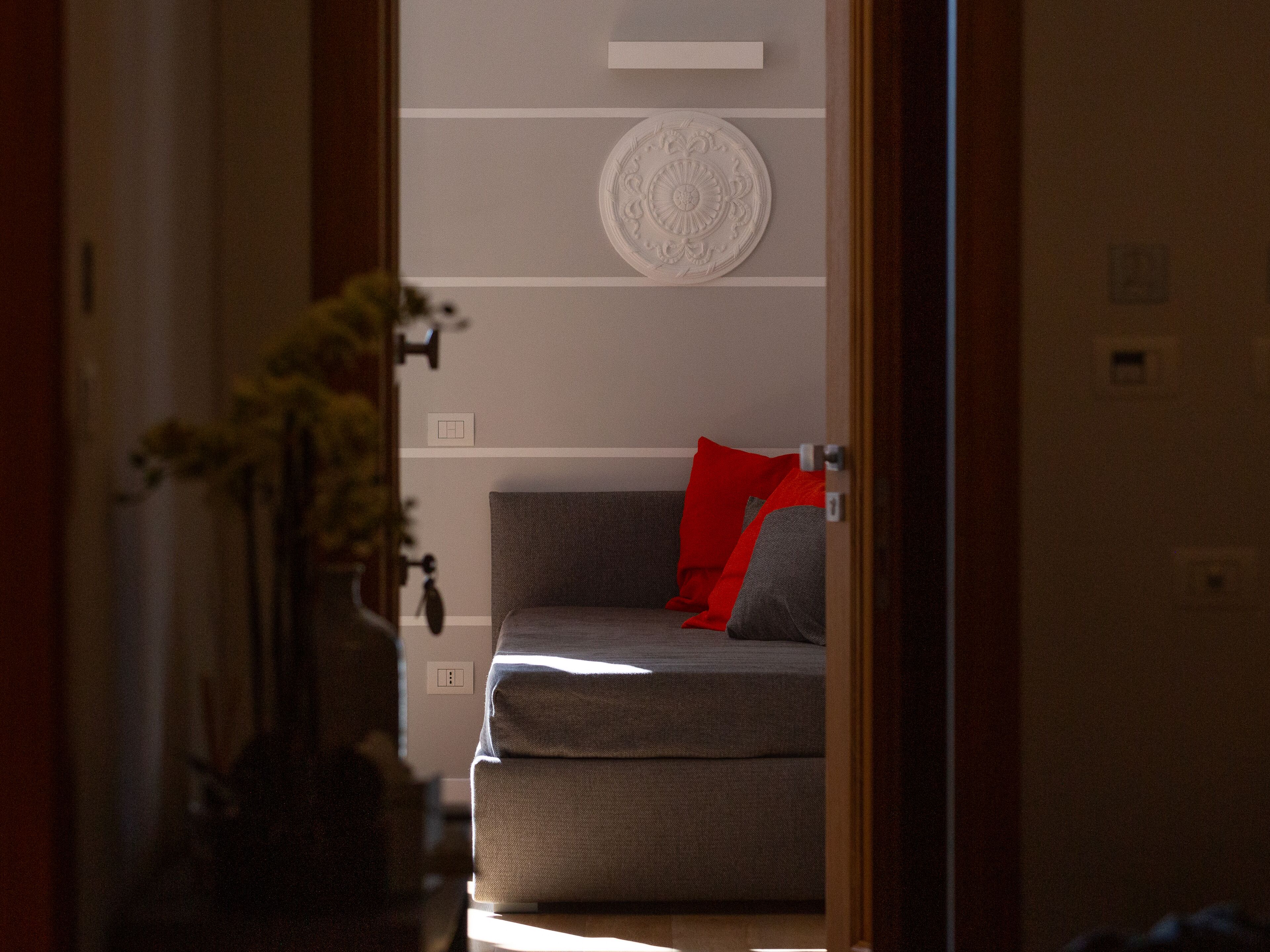 A room with a grey sofa, red and grey cushions, a decorative wall plate, and a vase with flowers.