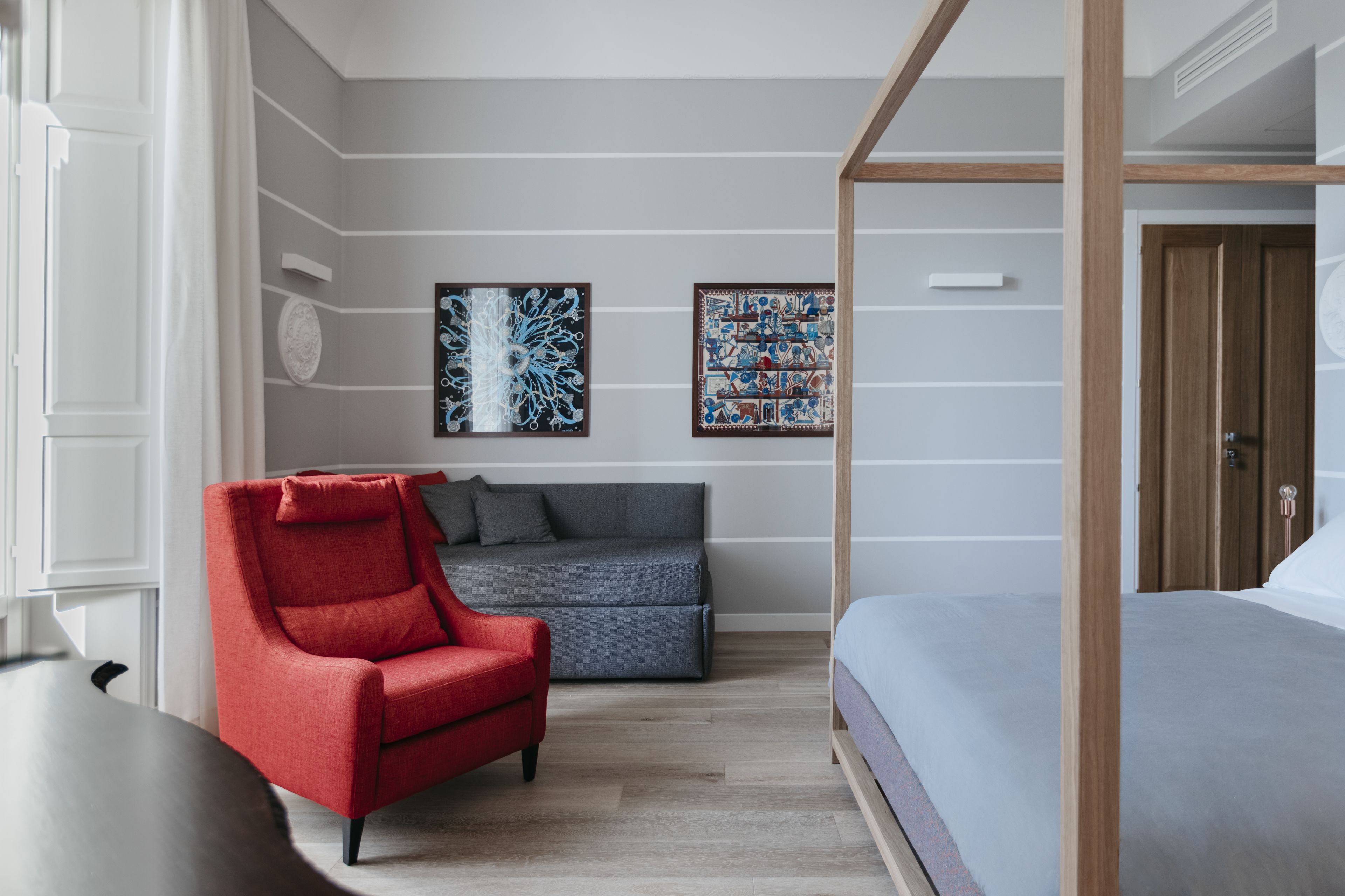 Um quarto moderno com uma cama de dossel, uma poltrona vermelha, um sofá cinzento e duas obras de arte na parede.