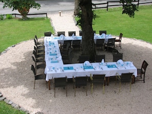 Un área de comedor al aire libre con mesas dispuestas para una comida, rodeada de sillas y un árbol.