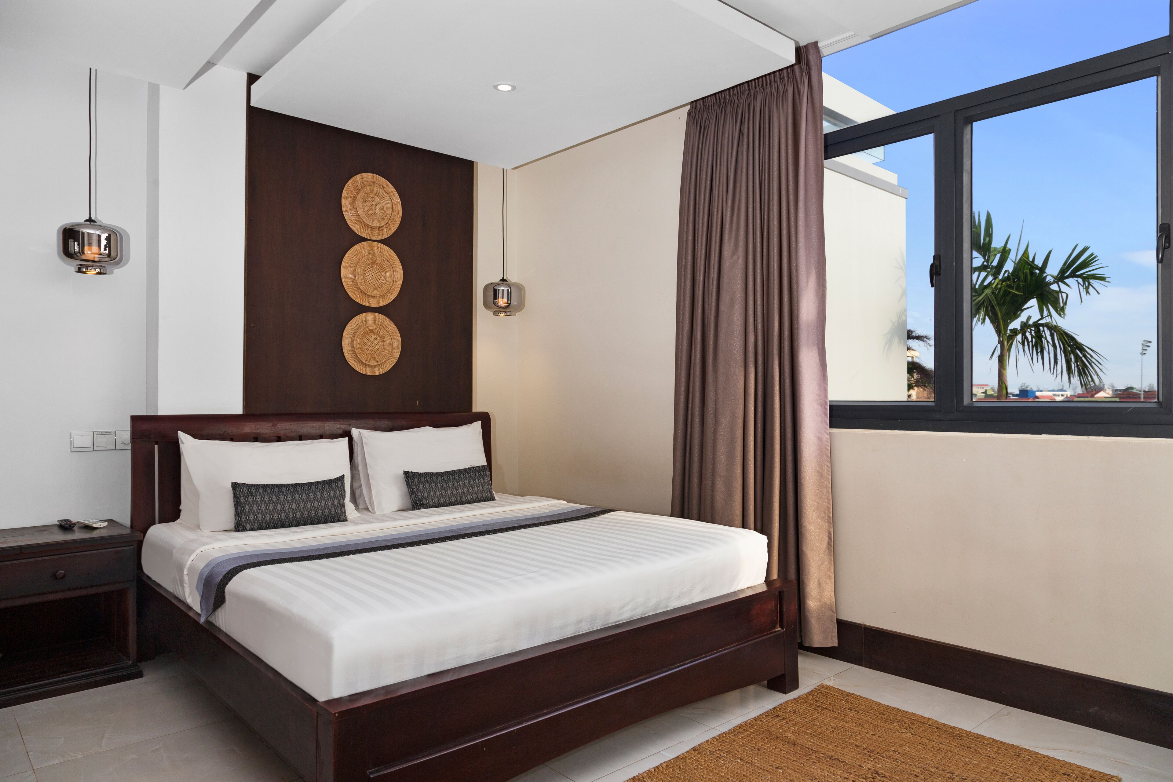A modern bedroom with a large bed, wooden headboard, and a view of palm trees through a window.