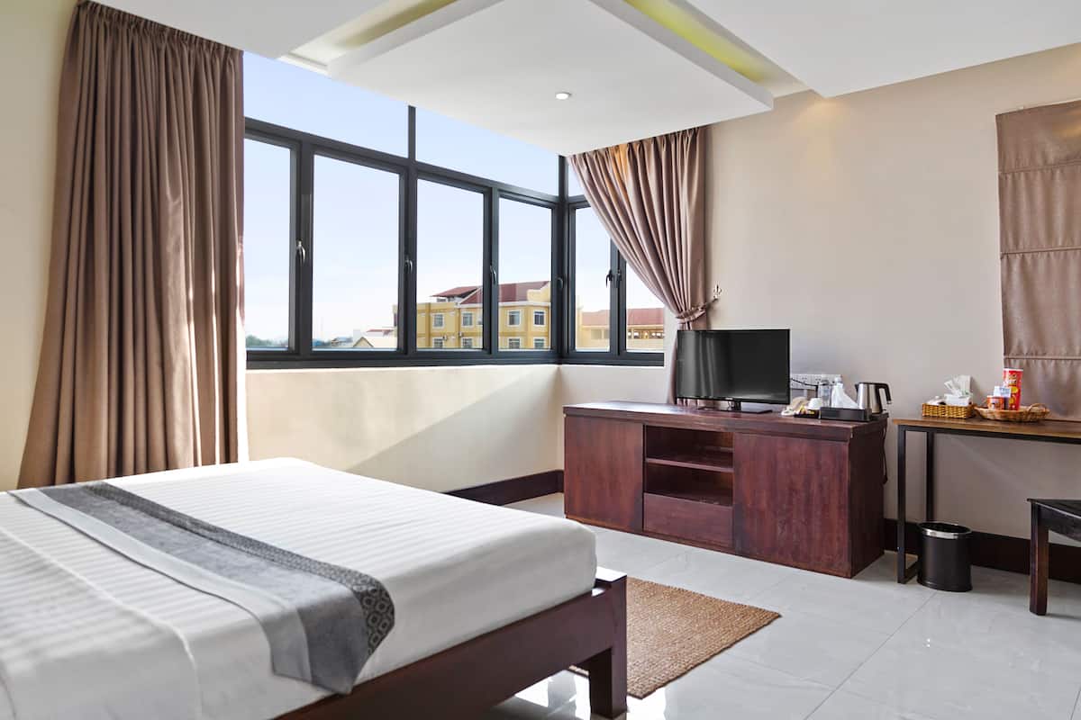 Habitación de hotel con una cama grande, un escritorio con televisor y vistas a edificios a través de la ventana.