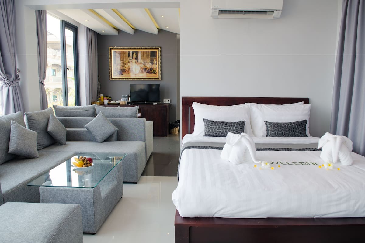 Una habitación de hotel moderna con un sofá gris, una mesa de centro de vidrio, una cama con ropa blanca y un cartel que dice 'BIENVENUE', además de un cuadro en la pared.