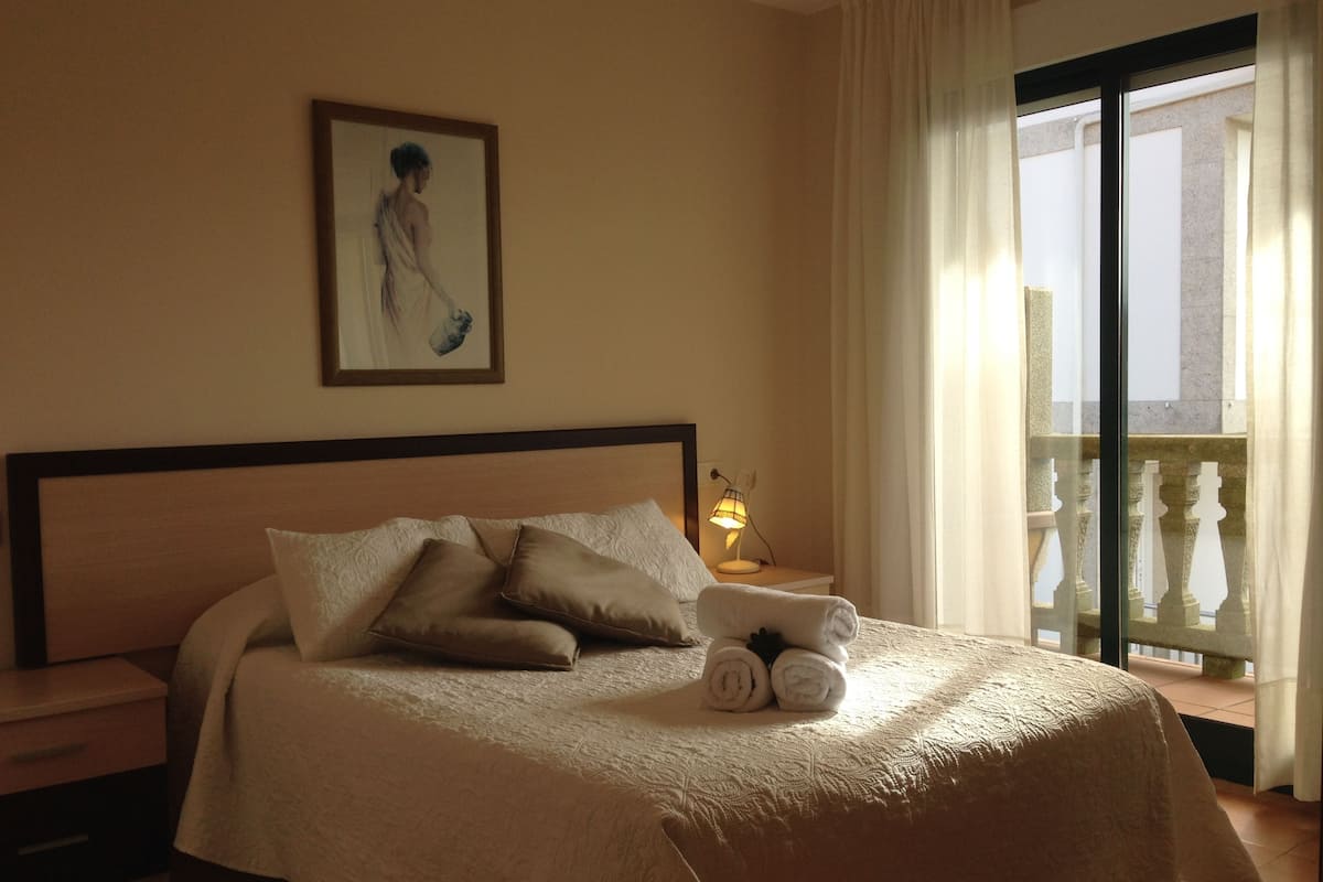 A hotel room with a bed, pillows, a nightstand with a lamp, a painting on the wall, and a balcony with a view.