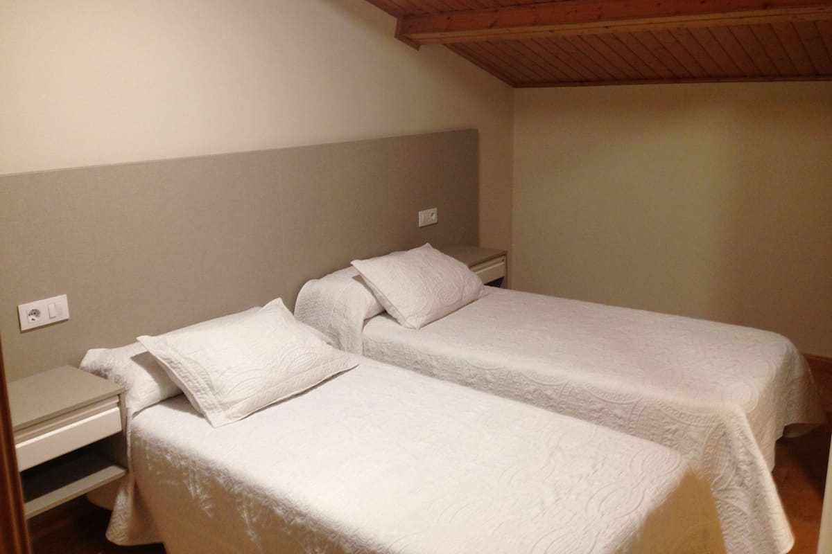 A room with two single beds, a wooden ceiling, and a plain wall with a light switch.