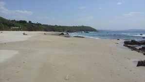 Ein weiter Sandstrand mit sanften Wellen, blauem Himmel und einer grünen Hügelkette im Hintergrund.