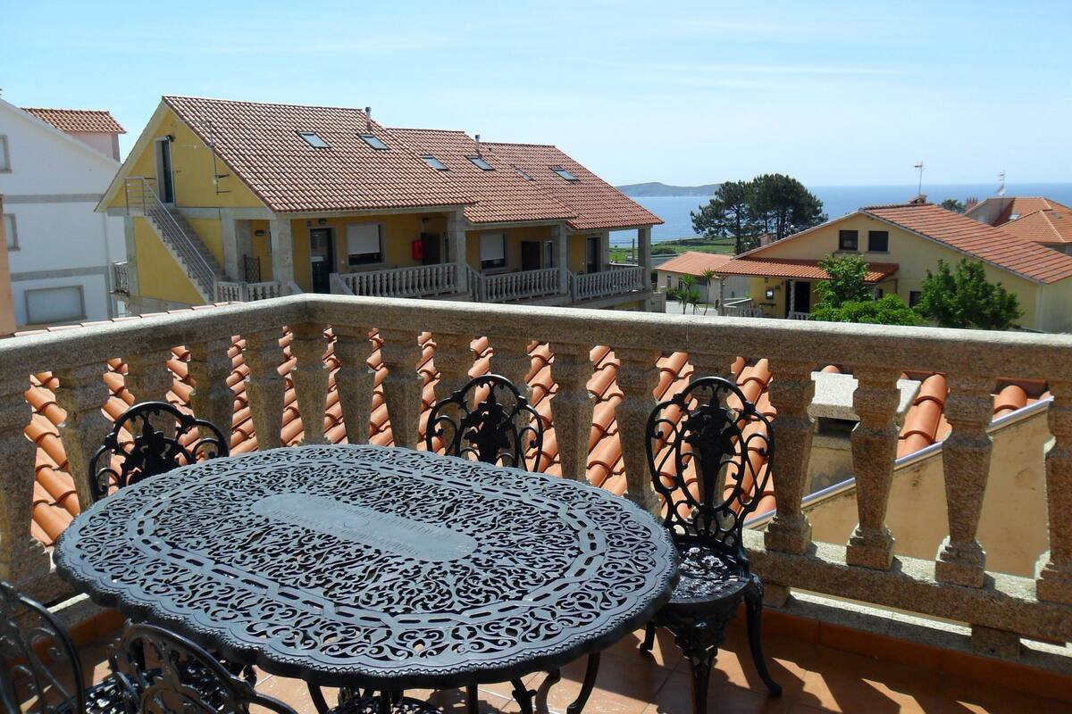 A balcony with a round metal table and chairs, overlooking a residential area with terracotta roofs and a clear sky.