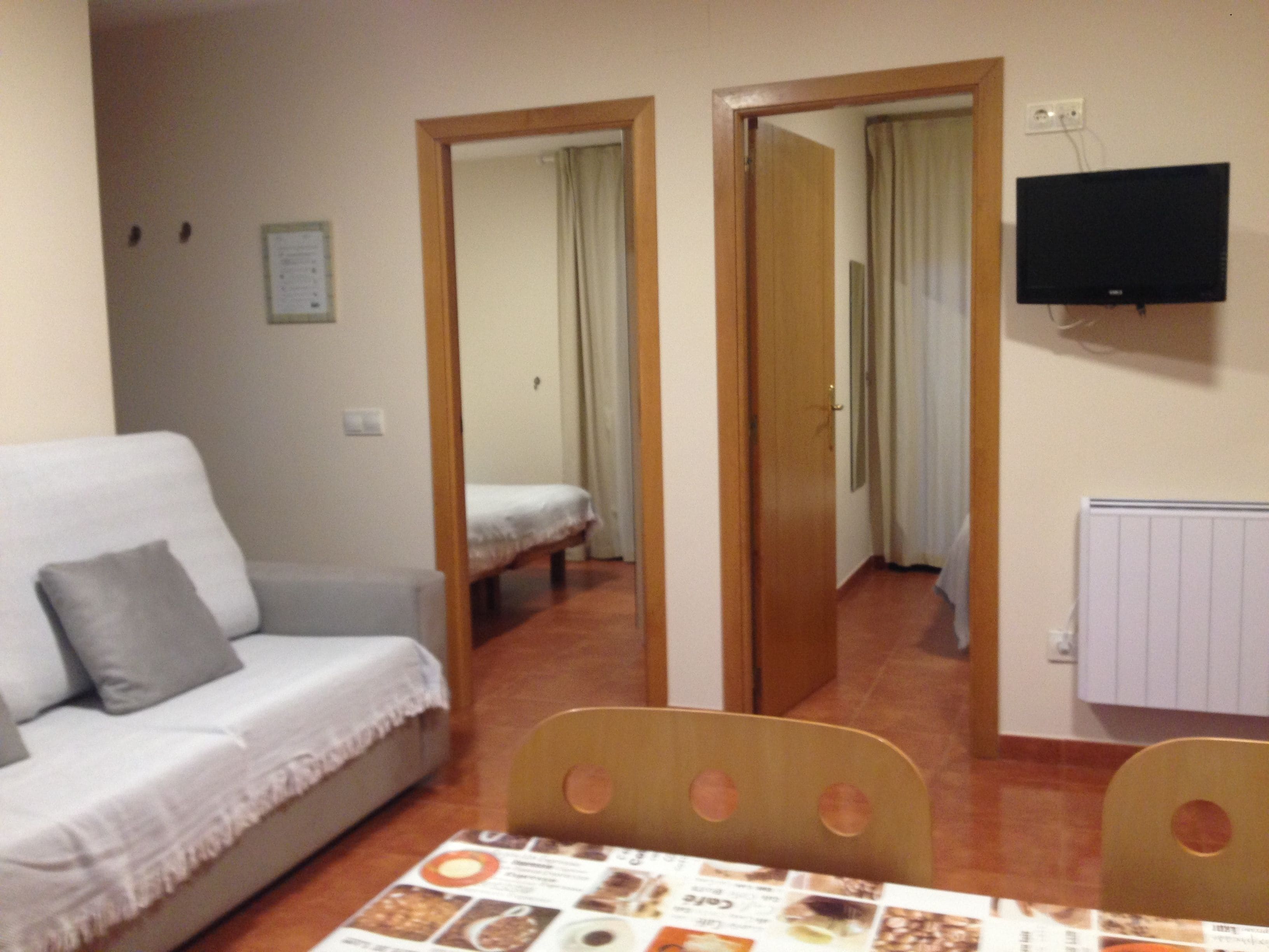 A room with a sofa, a dining table with chairs, a television mounted on the wall, and a door leading to another room.