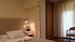 Ein Hotelzimmer mit zwei Betten, einem Nachttisch mit Lampe, einem Sessel und Blick auf ein fensterbedecktes Fenster.