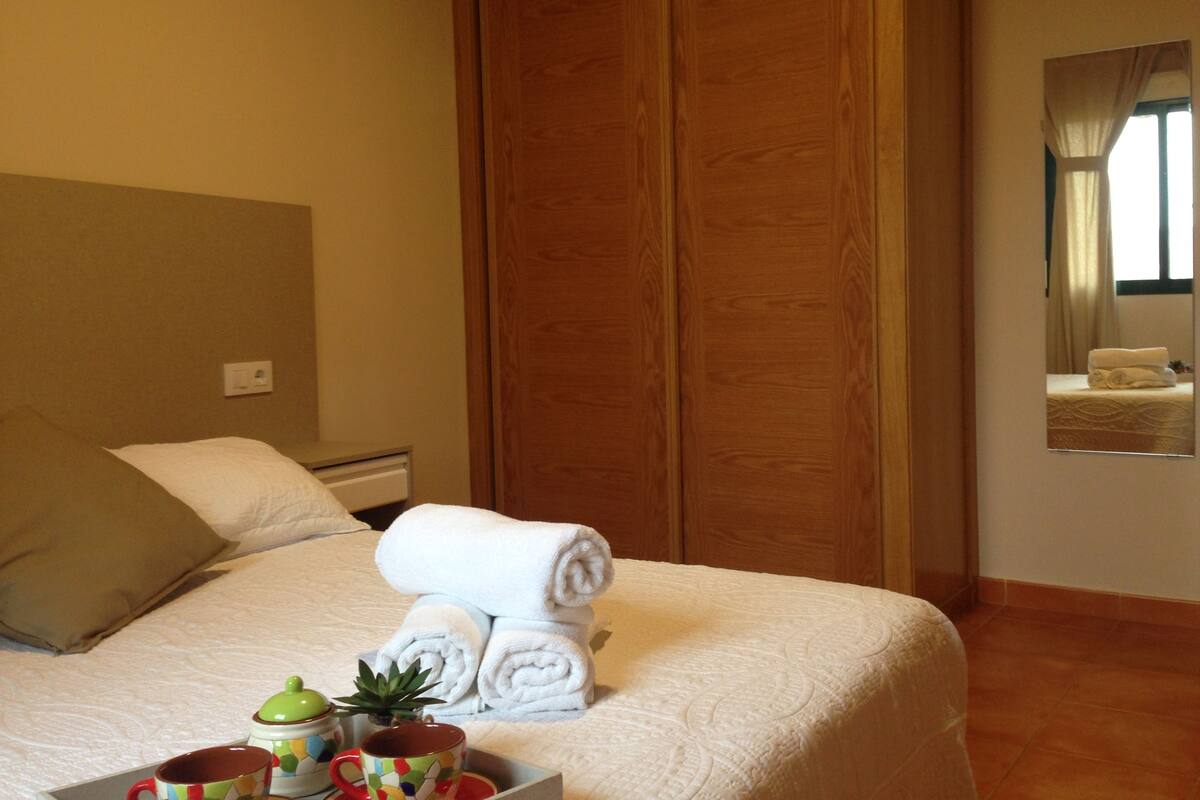 A neatly made bed with white linens, a bedside table with a cup and a small plant, and a wooden wardrobe in the background.