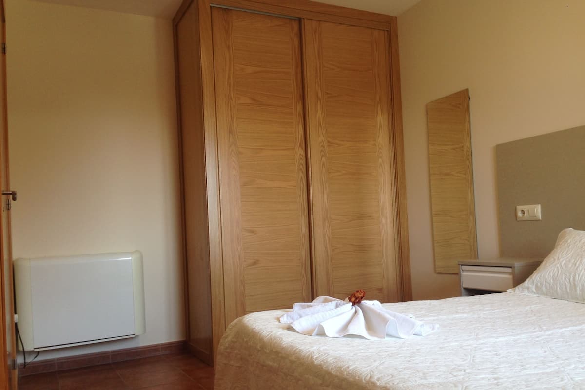 A bedroom with a large wooden wardrobe, a bed with a white bedspread, a bedside table, and a mirror on the wall.