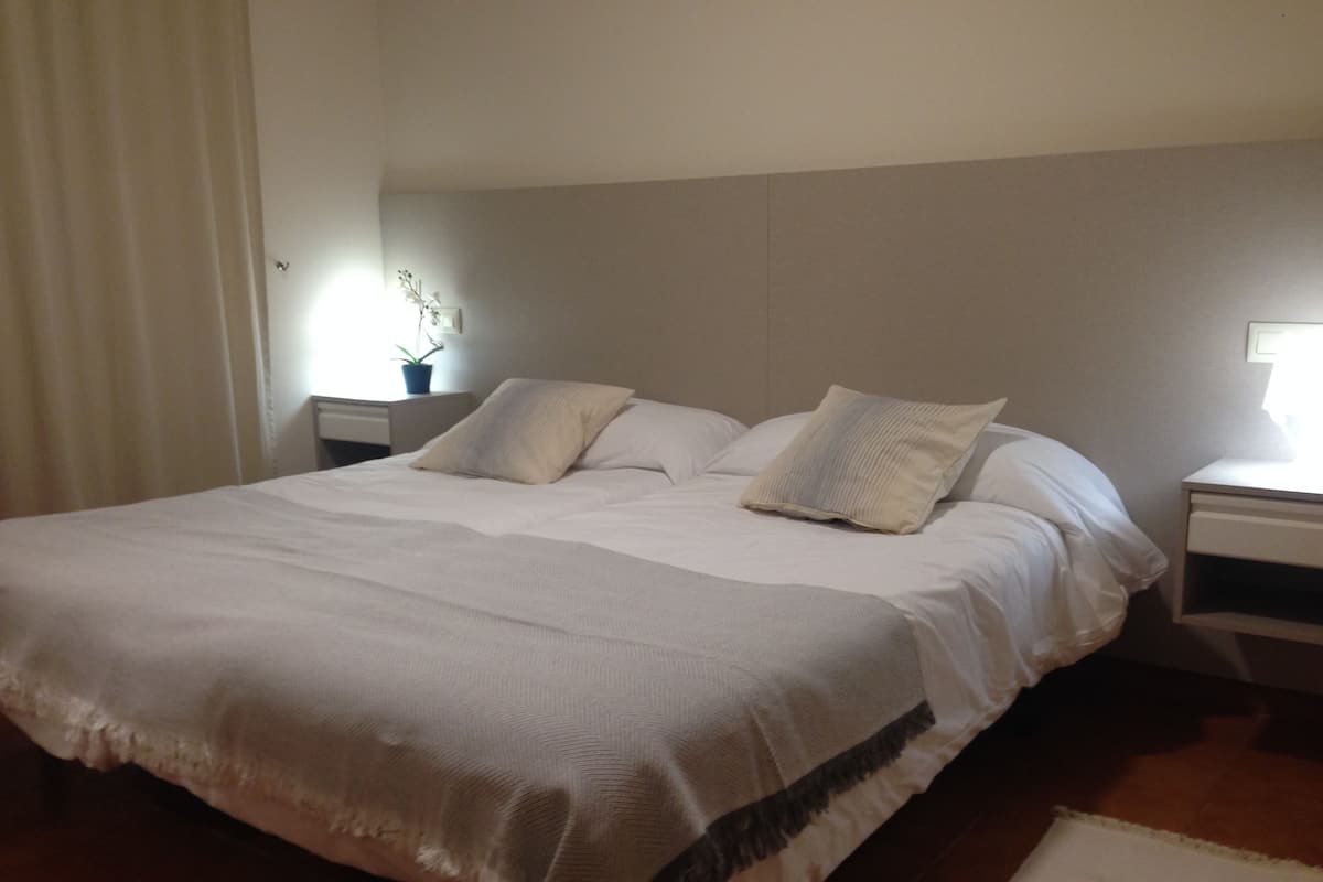 A neatly made bed with a white bedspread and pillows, a bedside table with a lamp, and a potted plant on the nightstand.