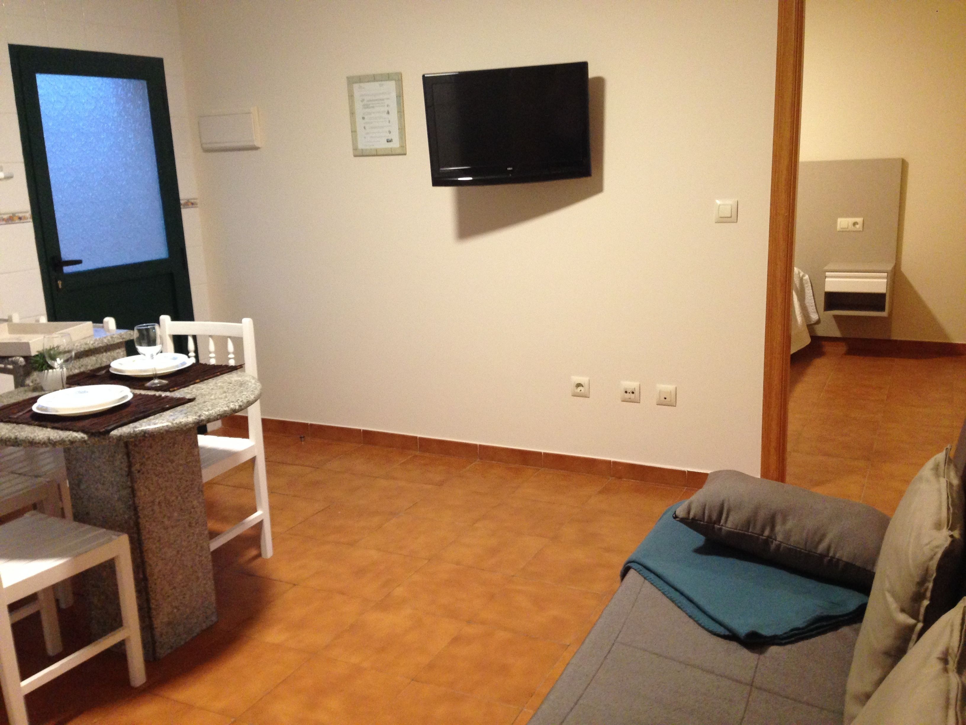 A small, well-lit room with a dining area featuring a table set for two, a television mounted on the wall, and a sofa with a blue throw pillow.