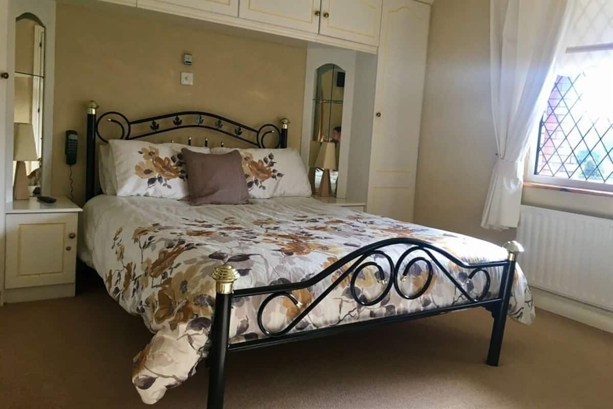 Een slaapkamer met een bed, nachtkastje, spiegel en raam met gordijnen.