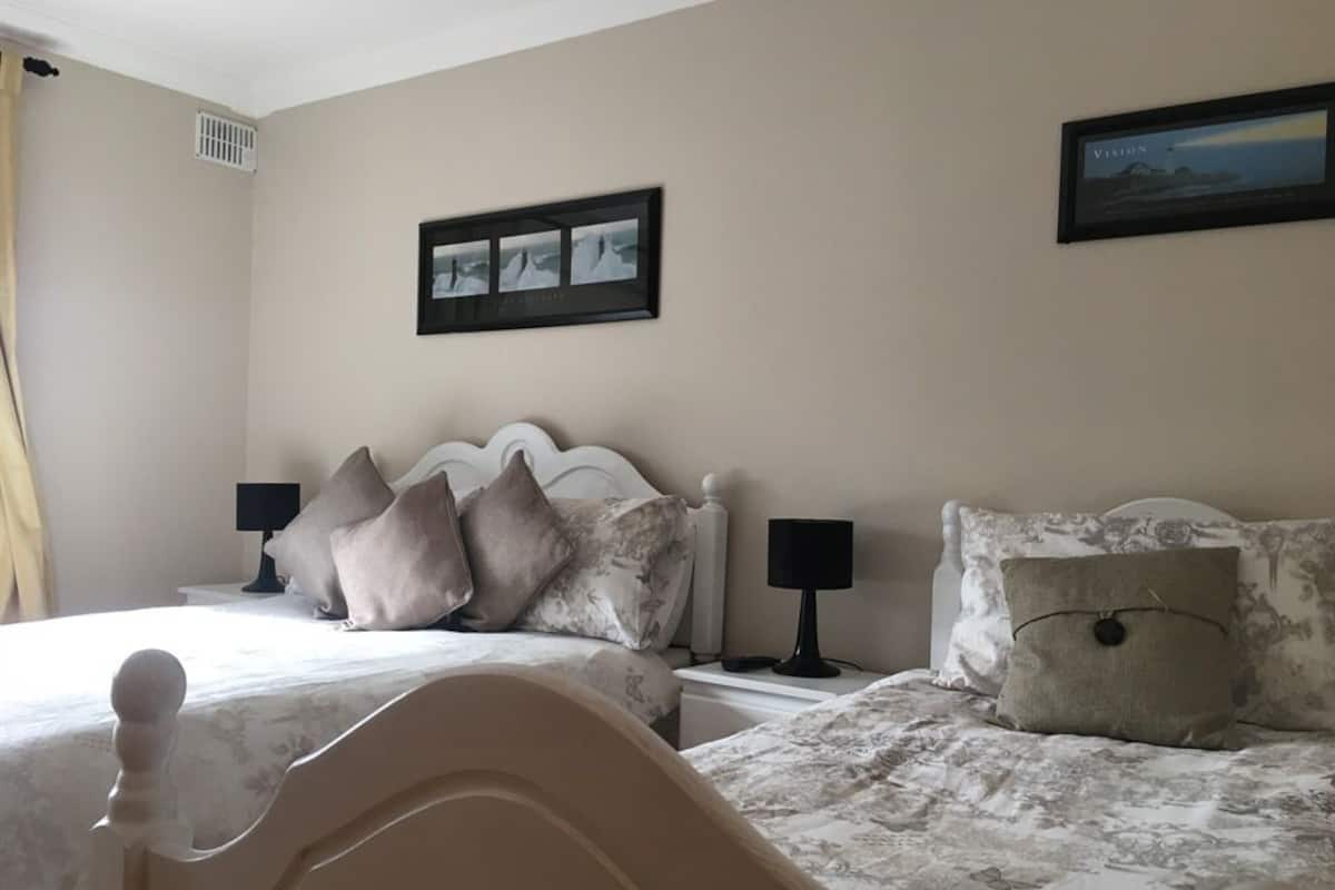 Een slaapkamer met twee bedden, elk met hoofdeinde, nachtkastjes, lampen en een lijst met een afbeelding aan de muur.