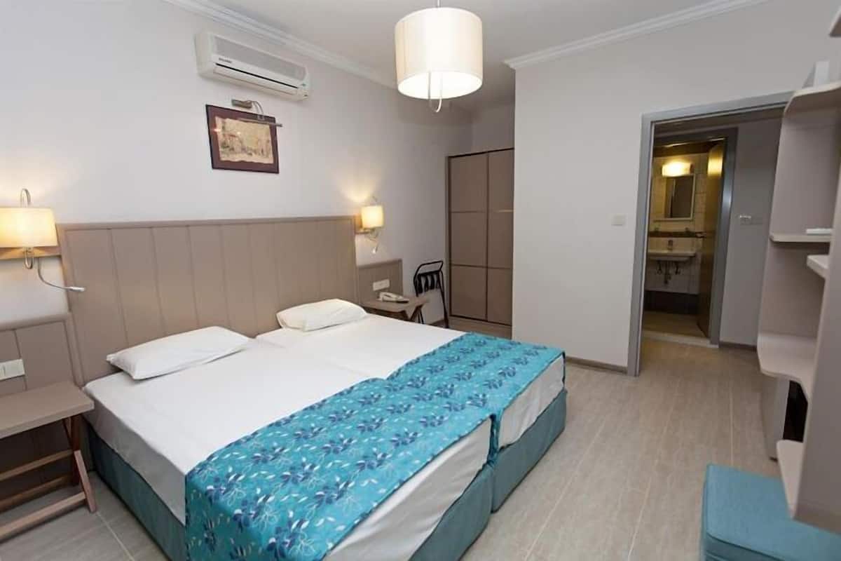 A hotel room with a bed, bedside tables, an air conditioning unit, a wall-mounted lamp, and a door leading to a bathroom.
