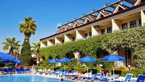 Un área de piscina con sillones de descanso y sombrillas, un edificio con balcón y una palmera.