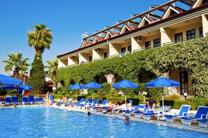 Un espace piscine avec des chaises longues et des parasols, un bâtiment doté d’un balcon et un palmier.