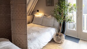 A cozy bedroom with a sloped ceiling, a bed with white bedding, a small table with a lamp, and a potted plant. - City House Grande Madame Agaath (Leeuwarden)