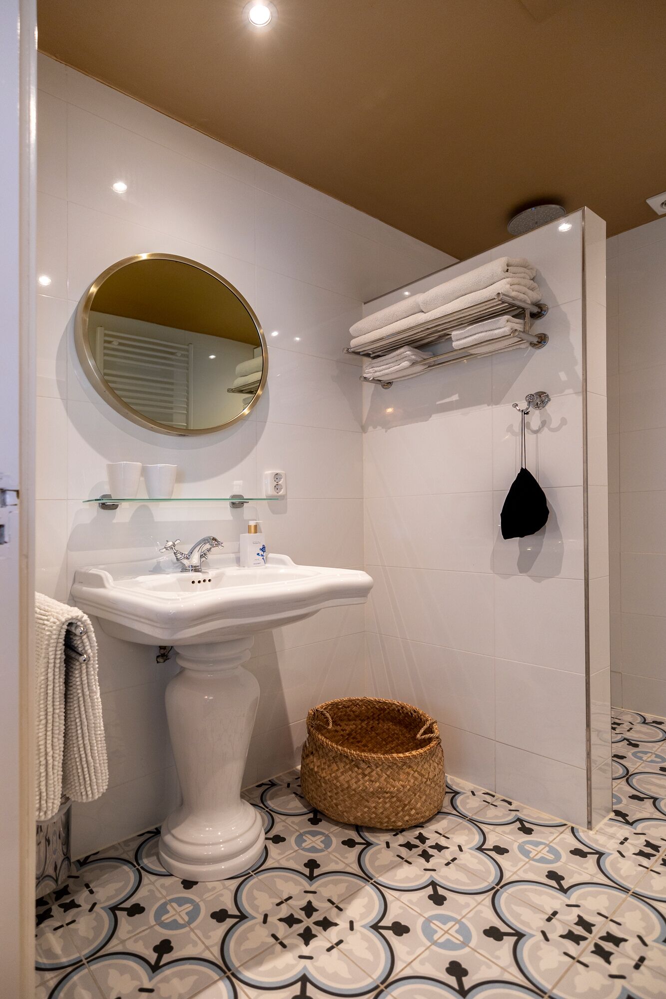 Un baño con lavamanos pedestal, espejo redondo, toallero, zona de ducha y una cesta tejida.