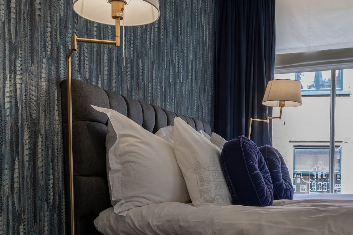 Une chambre avec un lit gris foncé, des oreillers blancs et bleus, deux lampes sur pied, un tabouret en bois et une fenêtre donnant sur la ville.