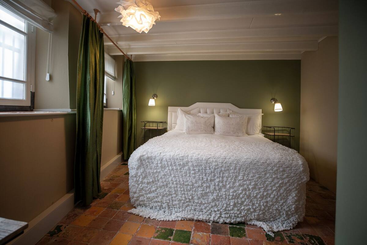 Un lit bien fait, recouvert d’une courtepointe blanche à motifs, avec deux lampes de chevet et un rideau vert.