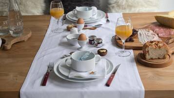 Ein gedeckter Frühstückstisch für zwei Personen, mit Tellern, Tassen, Besteck und verschiedenen Speisen wie Eiern, Brot und einer Obstschale.