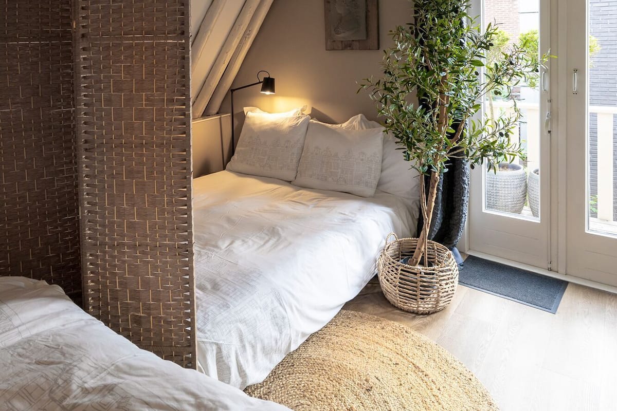 Une chambre avec un plafond en pente, un lit avec une literie blanche, une lampe de chevet, une plante en pot et une vue à travers une porte.
