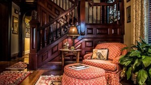 Una imponente escalera con barandillas de madera y un candelabro, que conduce a una habitación con un sofá otomano rojo con estampado, una planta verde grande y una alfombra estampada.