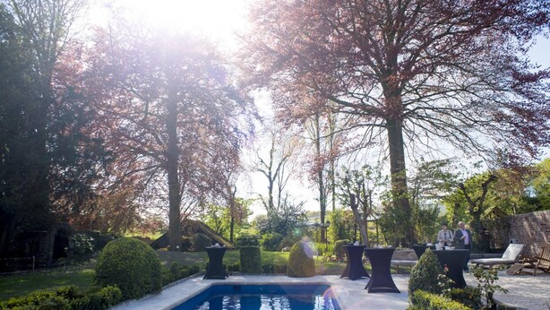 A swimming pool surrounded by neatly trimmed hedges and trees, with outdoor seating areas and a stone wall in the background. - B&B Filemon & Baucis (Bruges)